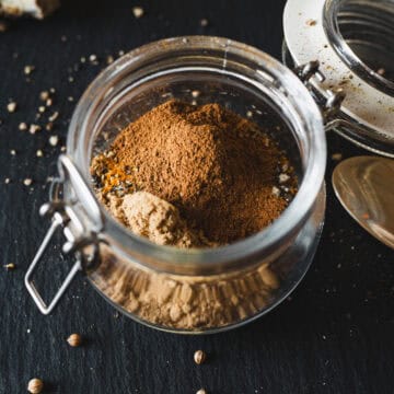 Open glass jar filled with fragrant Shawarma Spice, sitting on a dark surface with scattered spice seeds nearby.