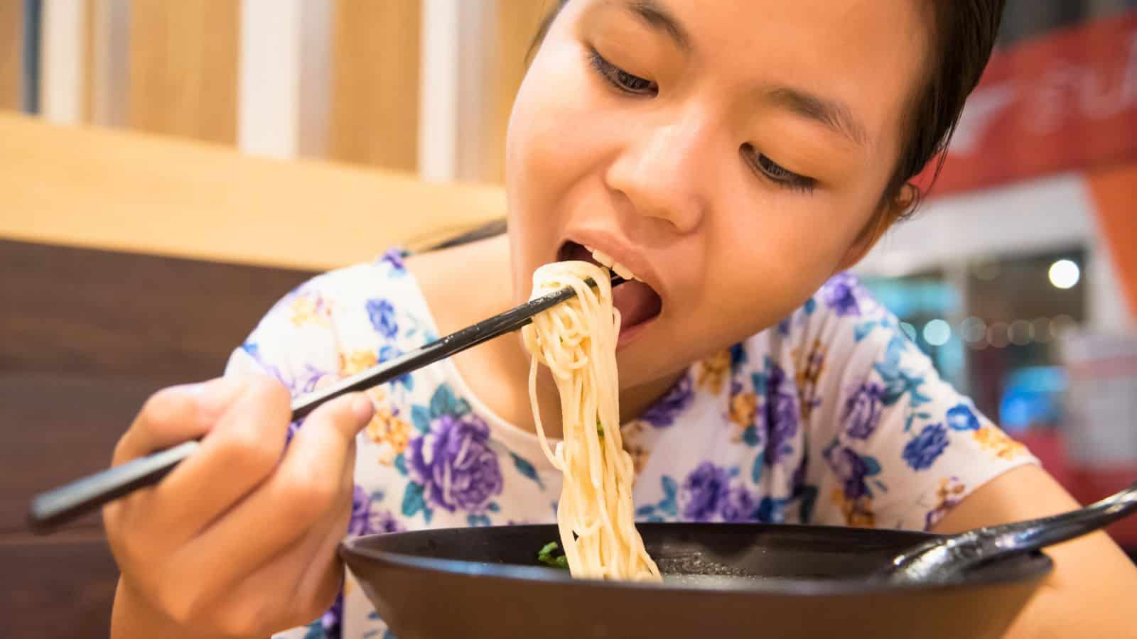 A person eating noodles with chopsticks from a black bowl at a restaurant.