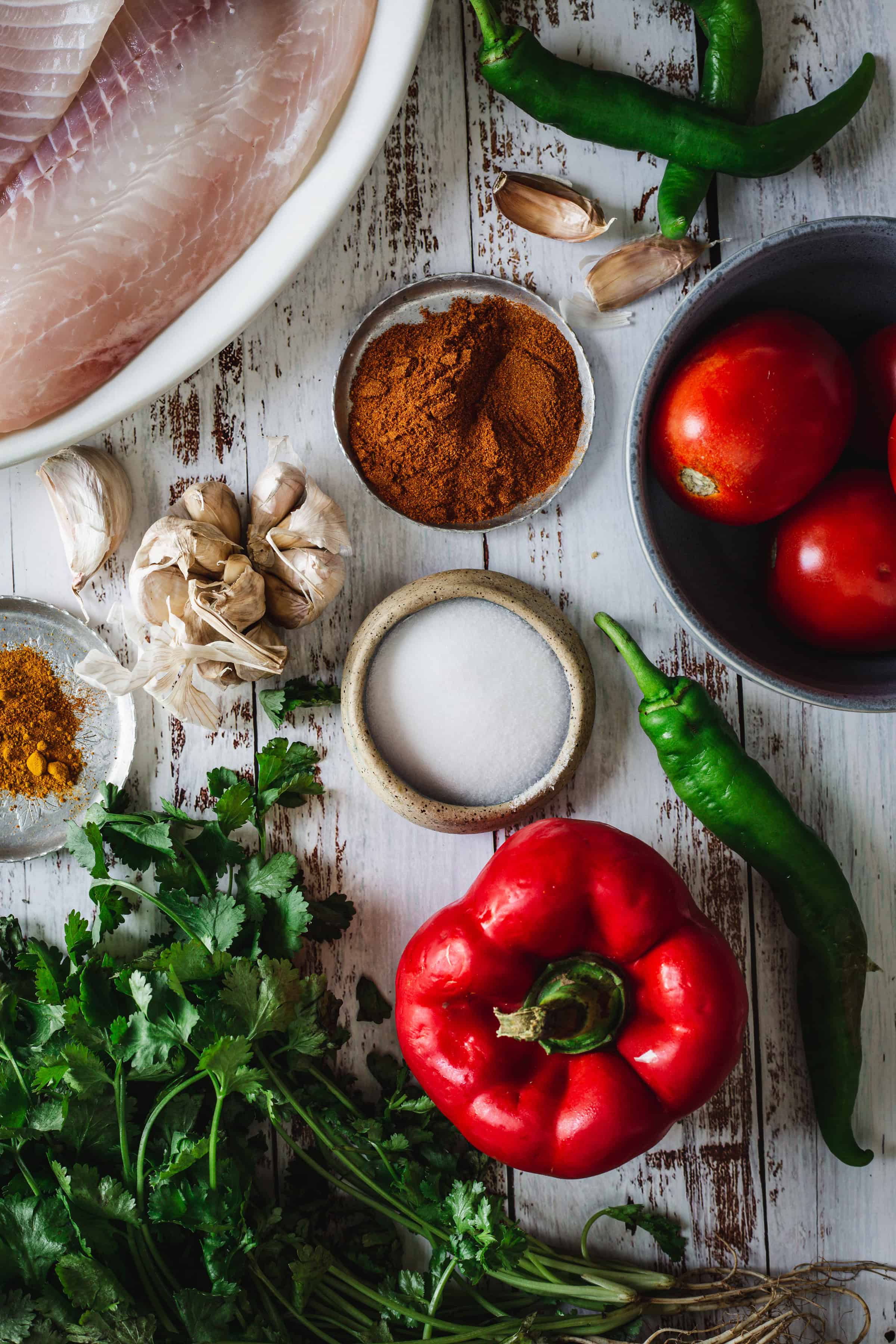 Fresh fish, tomatoes, garlic, cilantro, spices, green chilies, and a red bell pepper on a rustic white table.