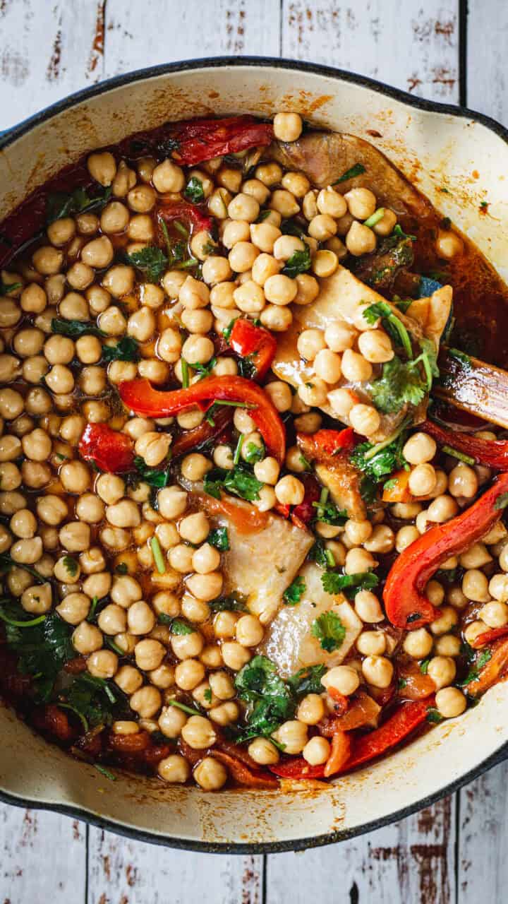 A pot of chickpea stew with red peppers, greens, and herbs on a rustic white wooden surface.