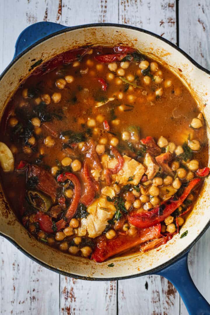 A blue pot filled with chickpea stew, vegetables, and spices on a rustic white wooden surface.