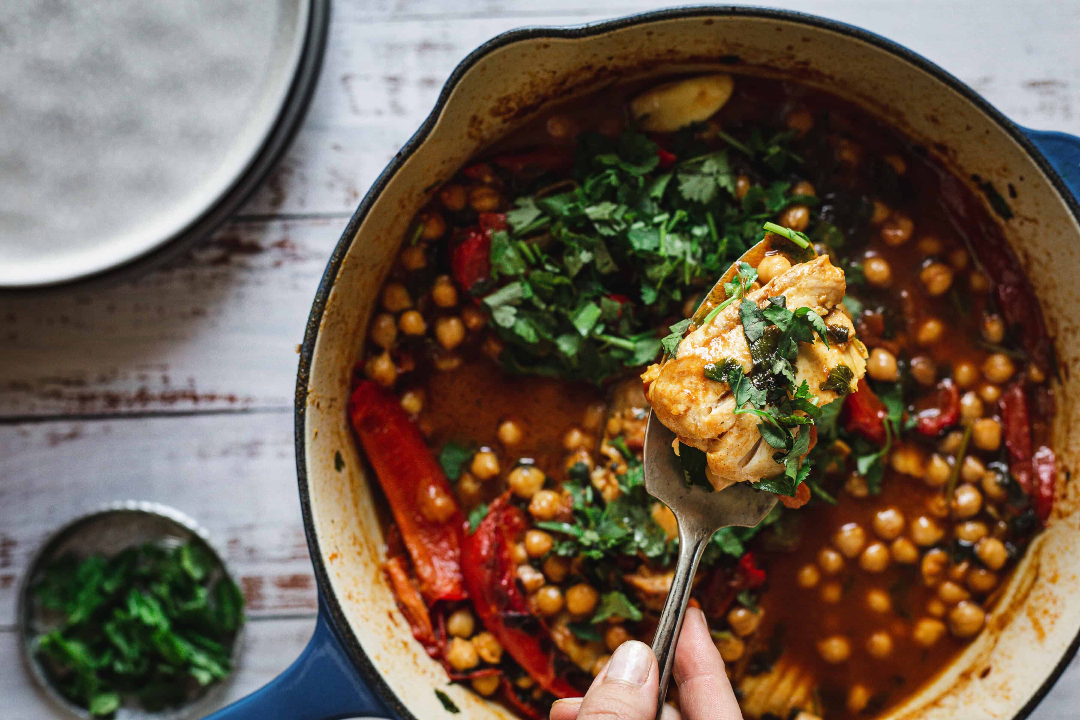 A hand holds a fork with chicken over a pot of chickpea stew, garnished with fresh herbs.