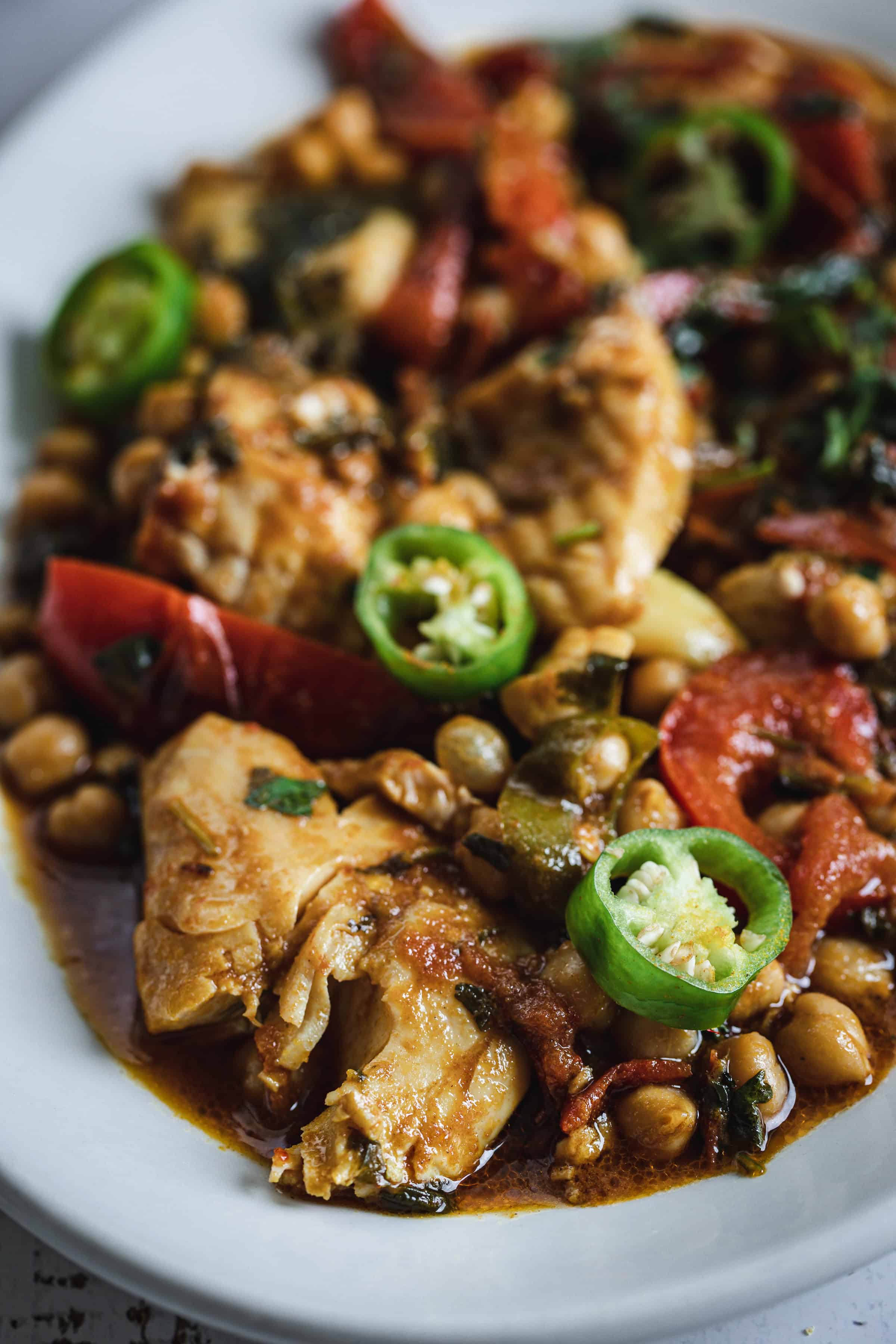 Chicken stew with chickpeas, tomatoes, herbs, and sliced jalapeños served on a white plate.