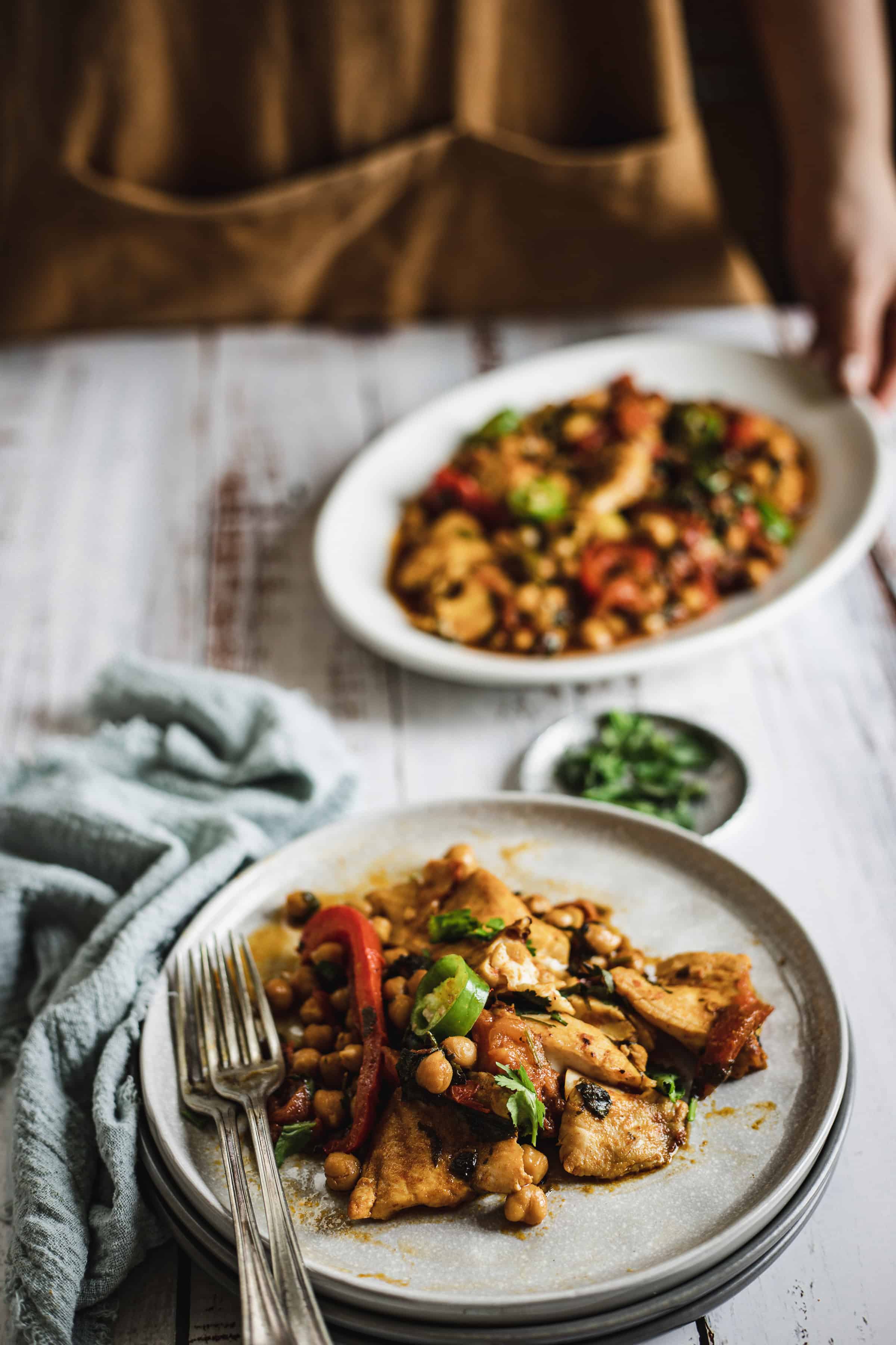 A plate of chickpea and vegetable stew with tofu, with another serving in the background on a white wooden table.