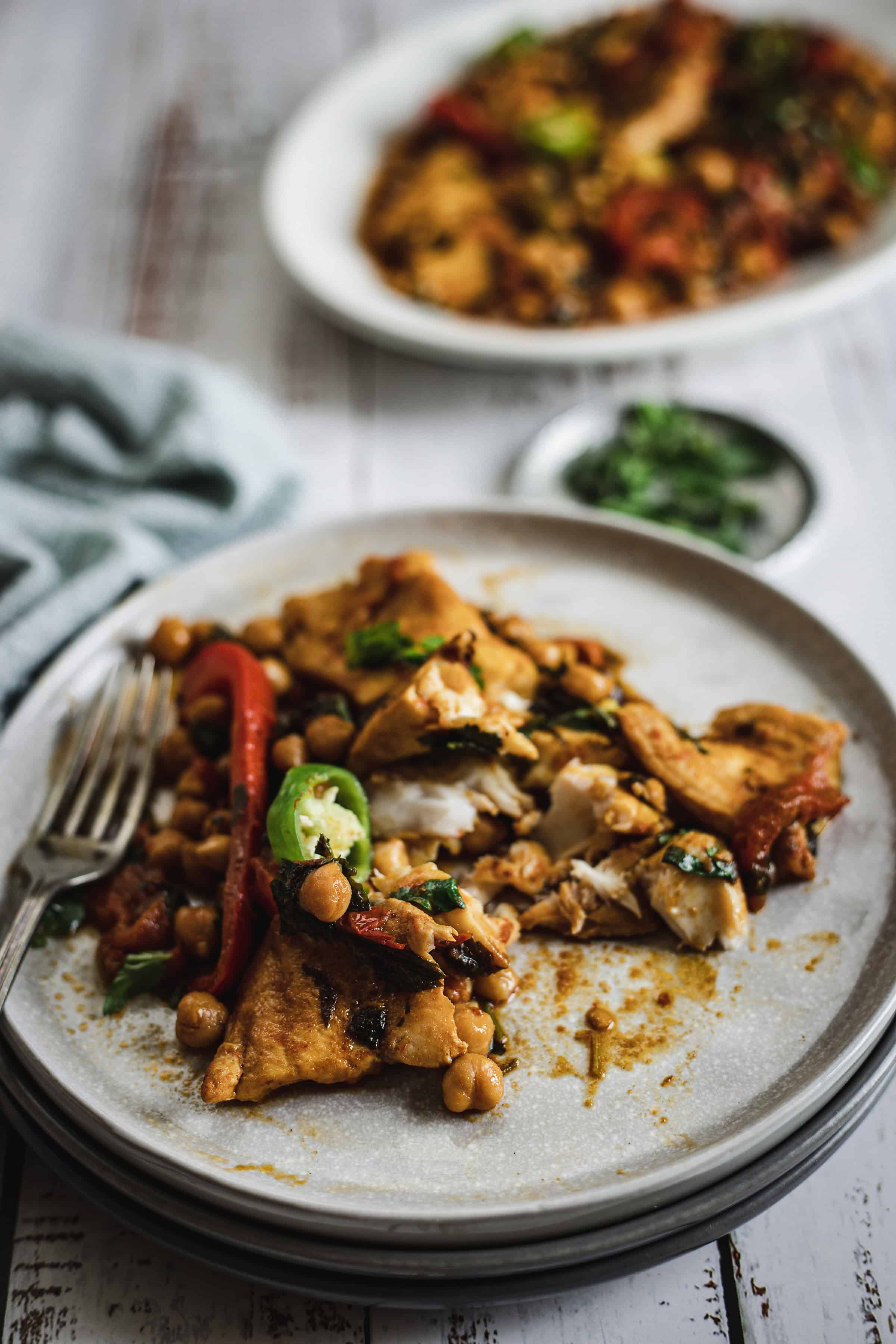 A plate of fish with chickpeas, red peppers, and herbs on a rustic white table.