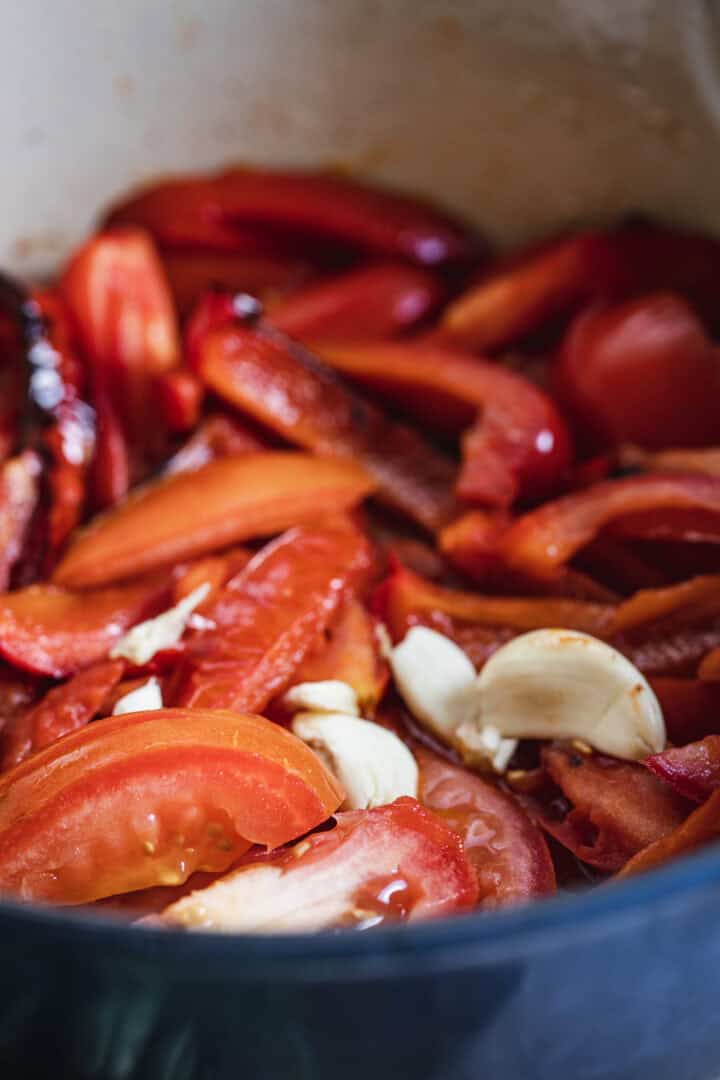 Sliced tomatoes, red peppers, and garlic cloves in a pot, ready for cooking.