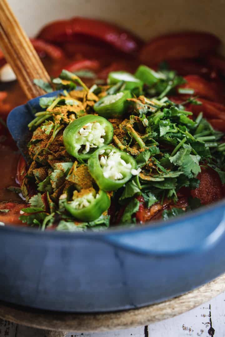 A blue pot with tomatoes, cilantro, spices, and sliced jalapeños being stirred with a wooden spoon.