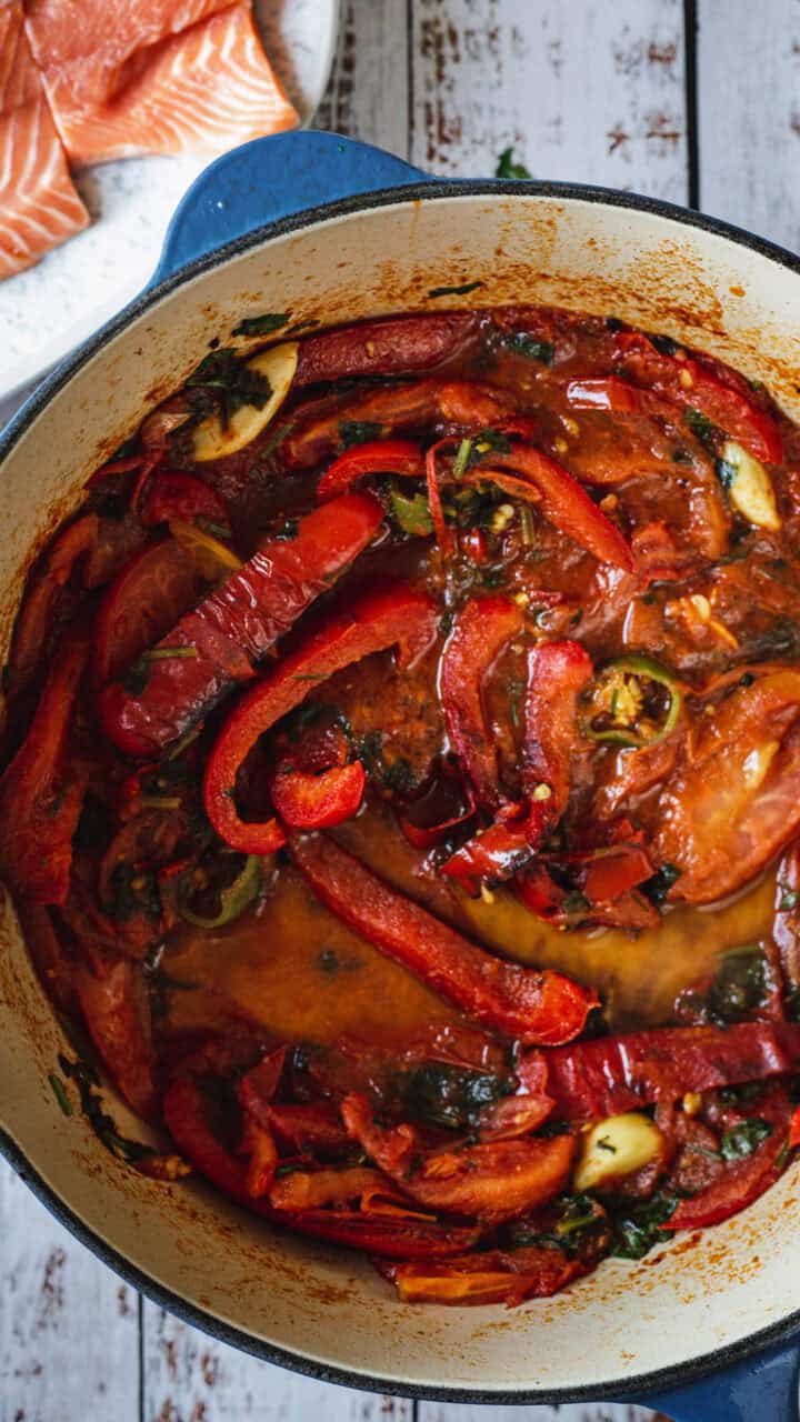 Red bell peppers, tomatoes, and greens simmer in a pot; raw salmon pieces are on a plate nearby.