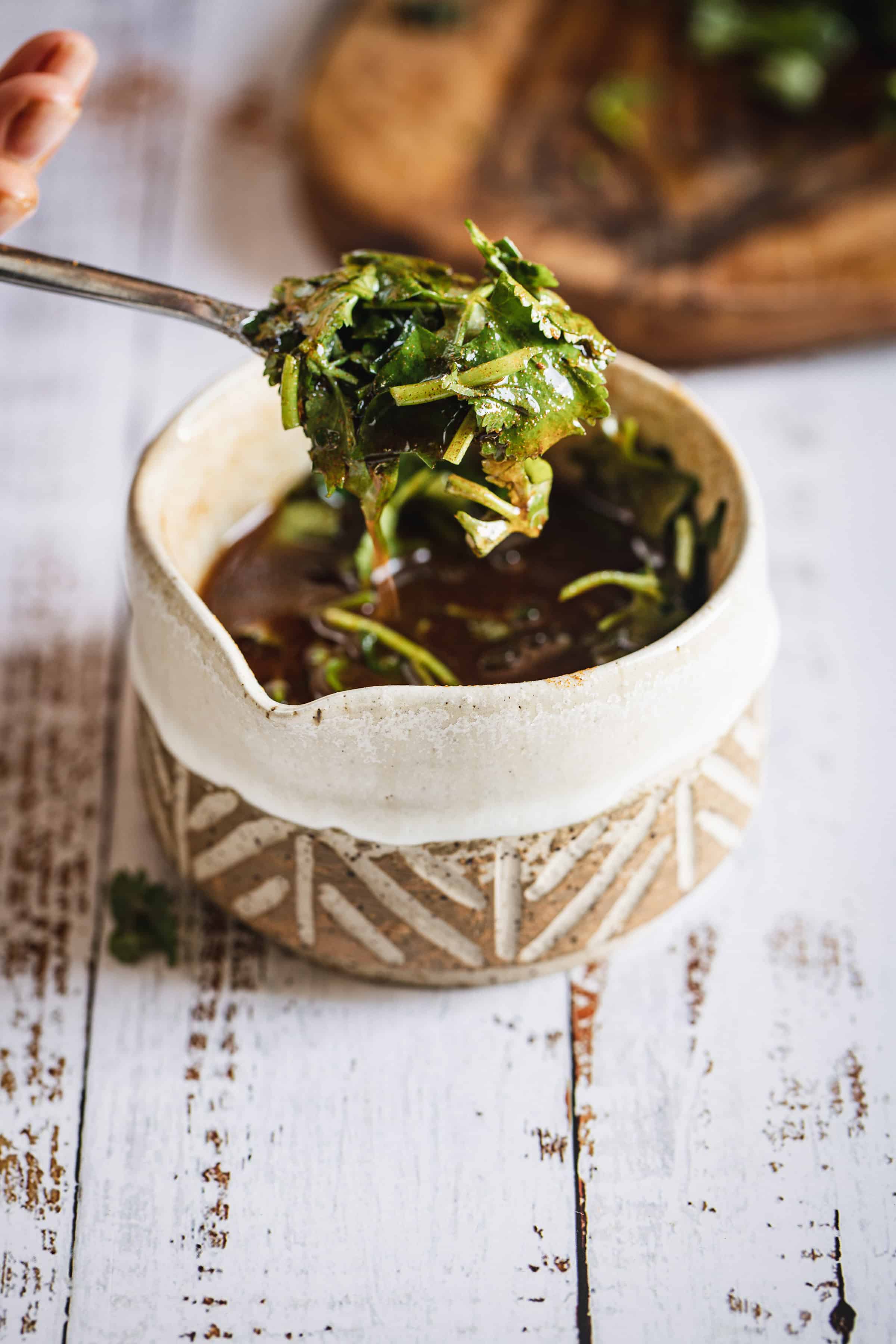 A spoon scoops fresh herbs from a bowl of dark broth on a rustic white wooden table.