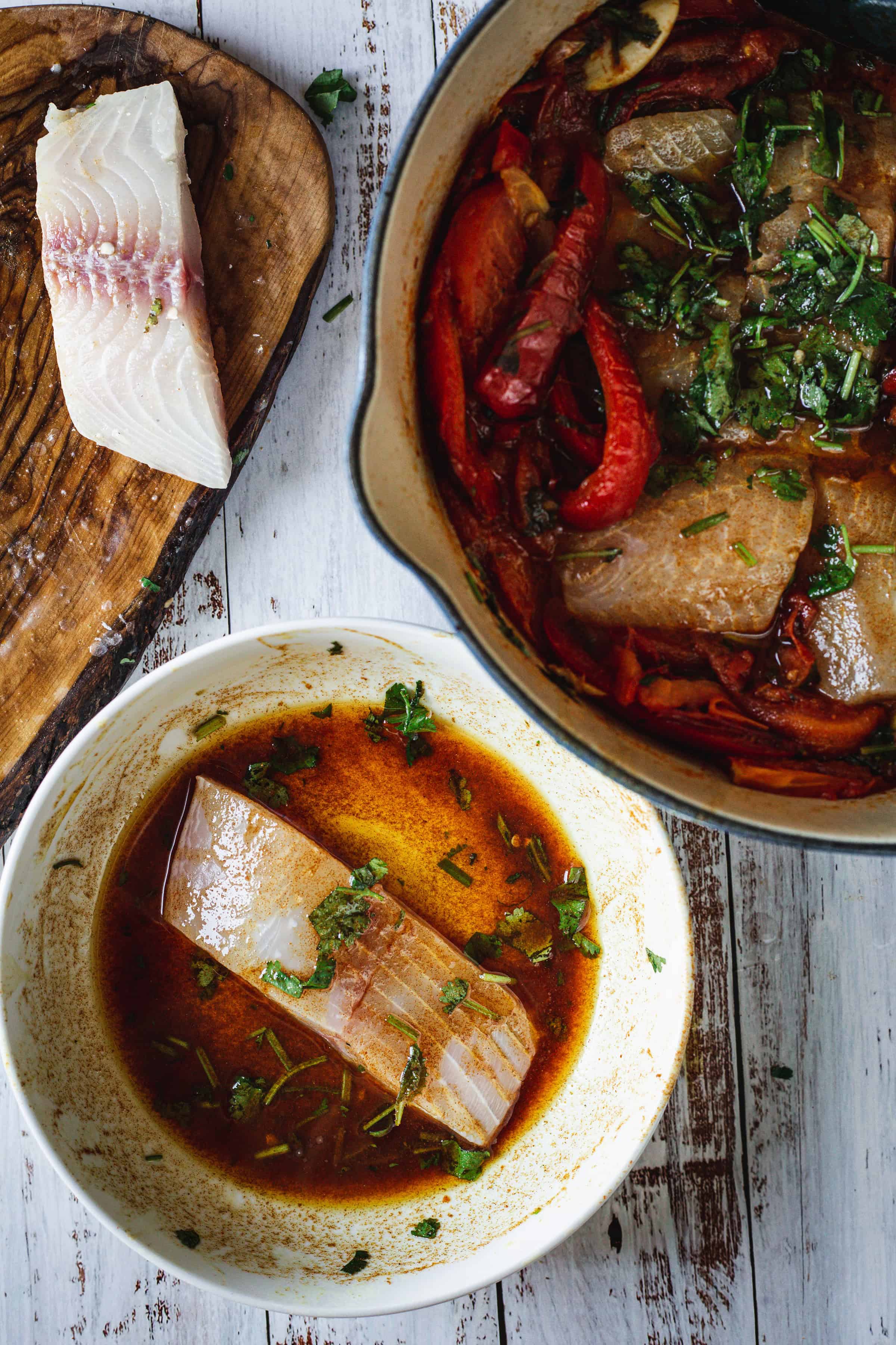 Raw fish in marinade, a pot of cooked fish with vegetables, and a fish fillet on a wooden board.