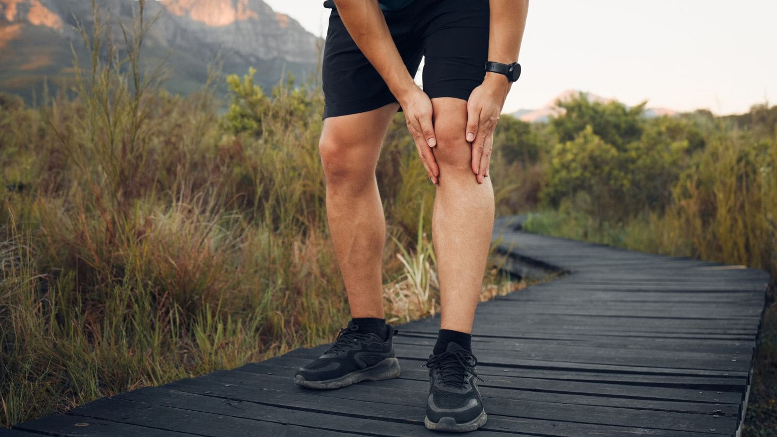 Person holding their knee in pain while standing on a wooden path outdoors, wearing athletic clothes&mdash;highlighting the importance of being aware of canning dangers and other potential outdoor risks.