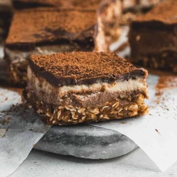 A chocolate oat dessert bars with creamy layers, dusted with cocoa powder, on parchment paper.