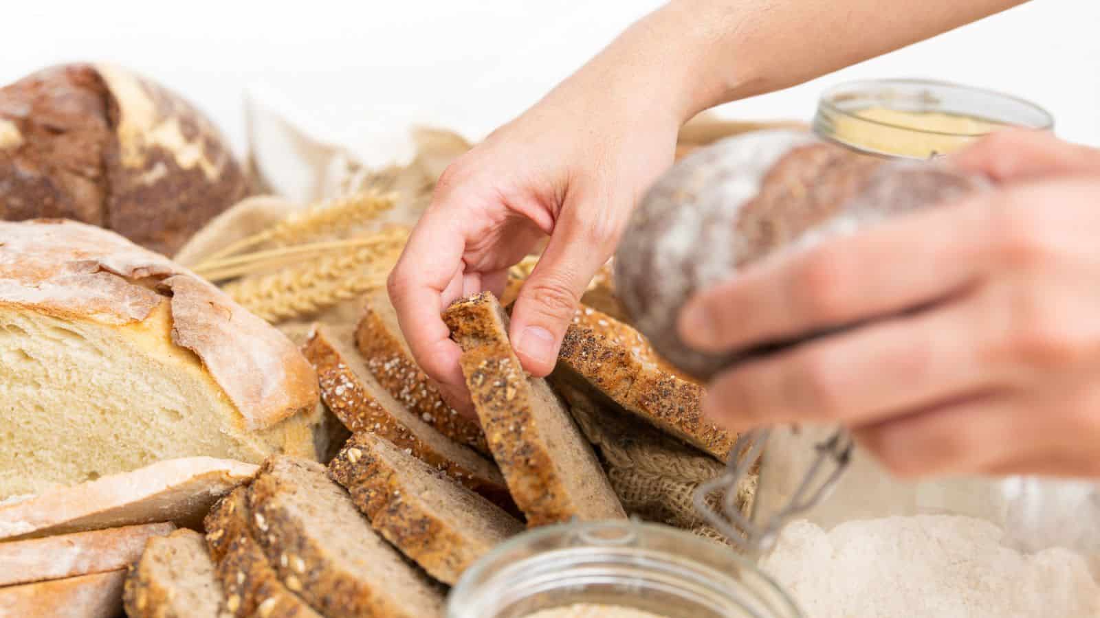 Hands picking up slices of multigrain bread from a variety of fresh, sliced loaves on a table.