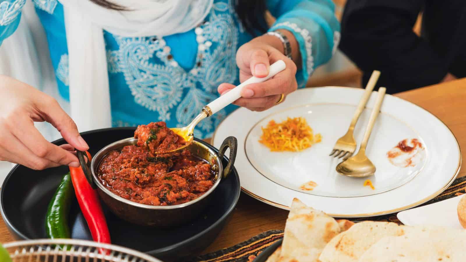A person in blue serves spicy curry from a bowl onto a plate beside naan bread and colorful chilies.