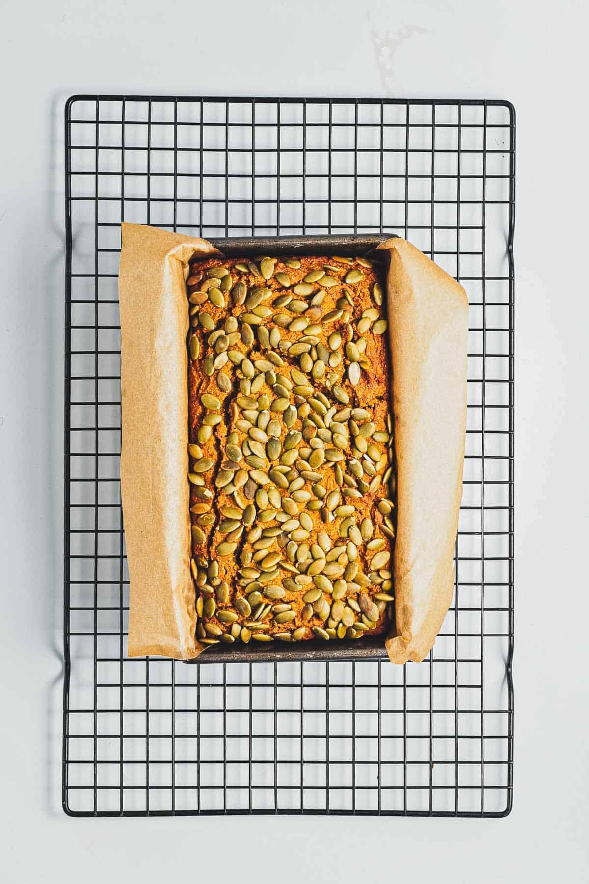 A loaf of bread topped with pumpkin seeds in a parchment-lined pan on a cooling rack.