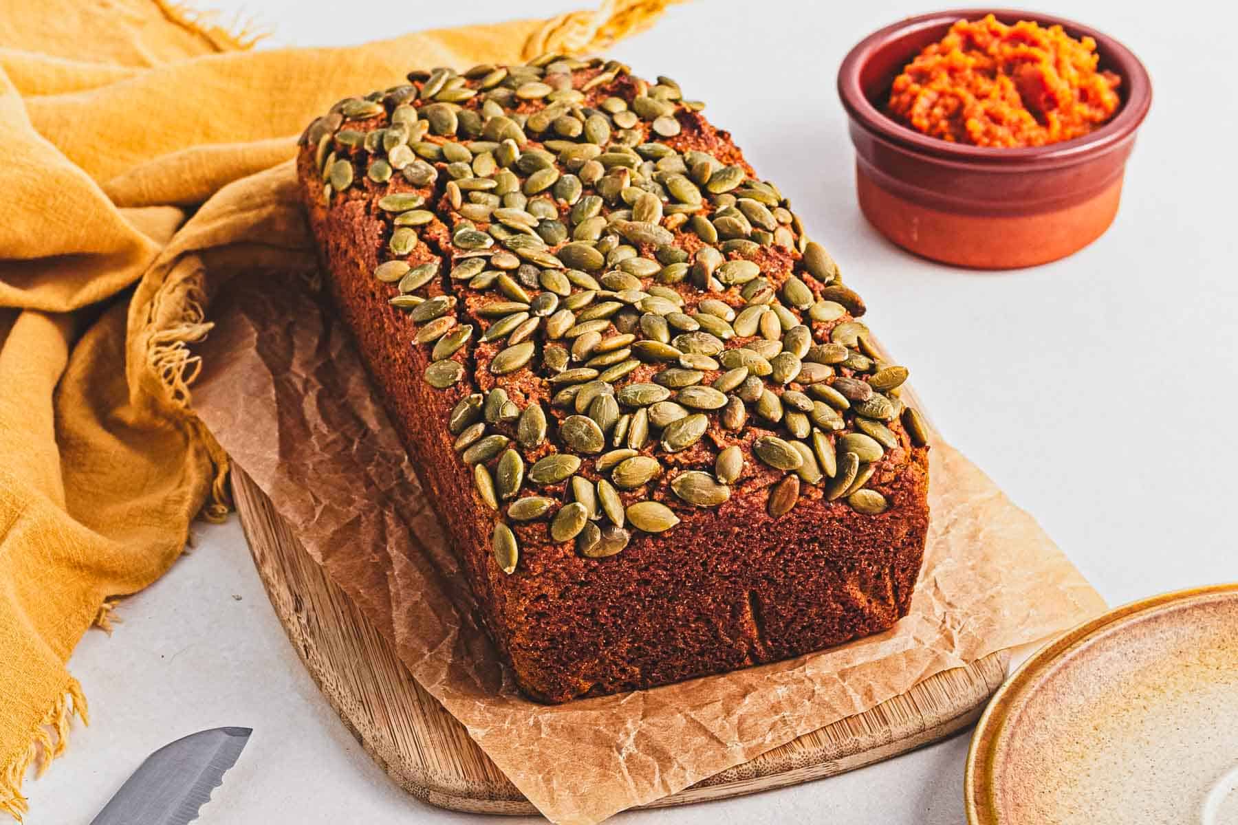 A loaf of pumpkin bread topped with pepitas on parchment, beside a bowl of pumpkin puree and a yellow cloth.