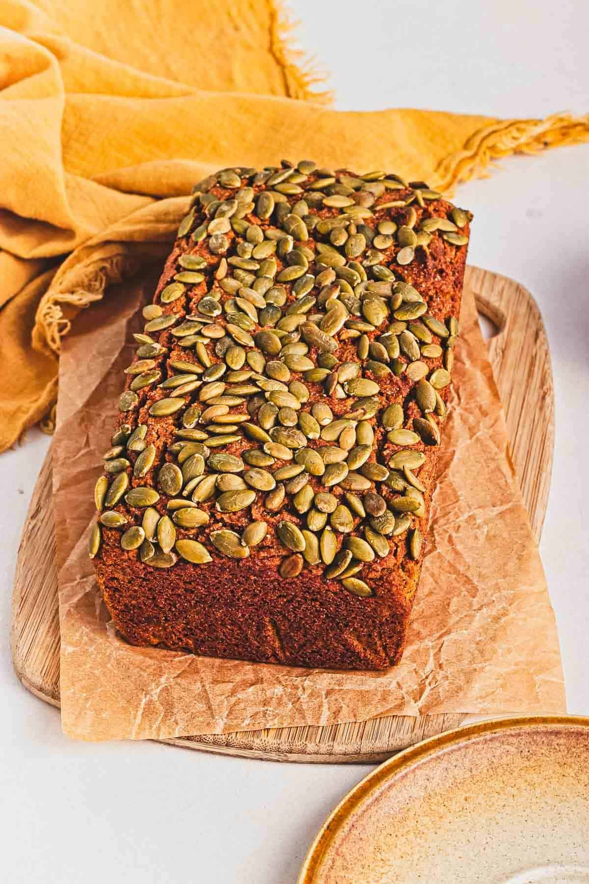 A loaf of bread topped with pumpkin seeds sits on parchment paper on a wooden board.