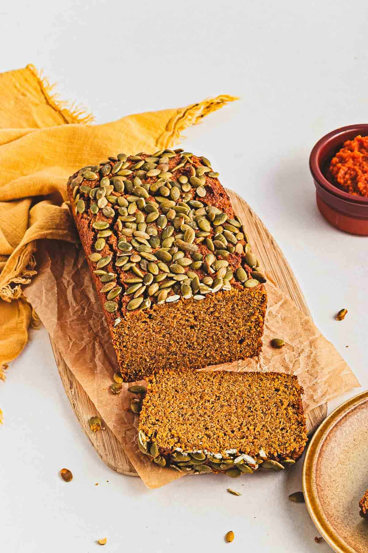 A sliced loaf of pumpkin bread topped with pumpkin seeds on parchment paper, next to a yellow cloth.