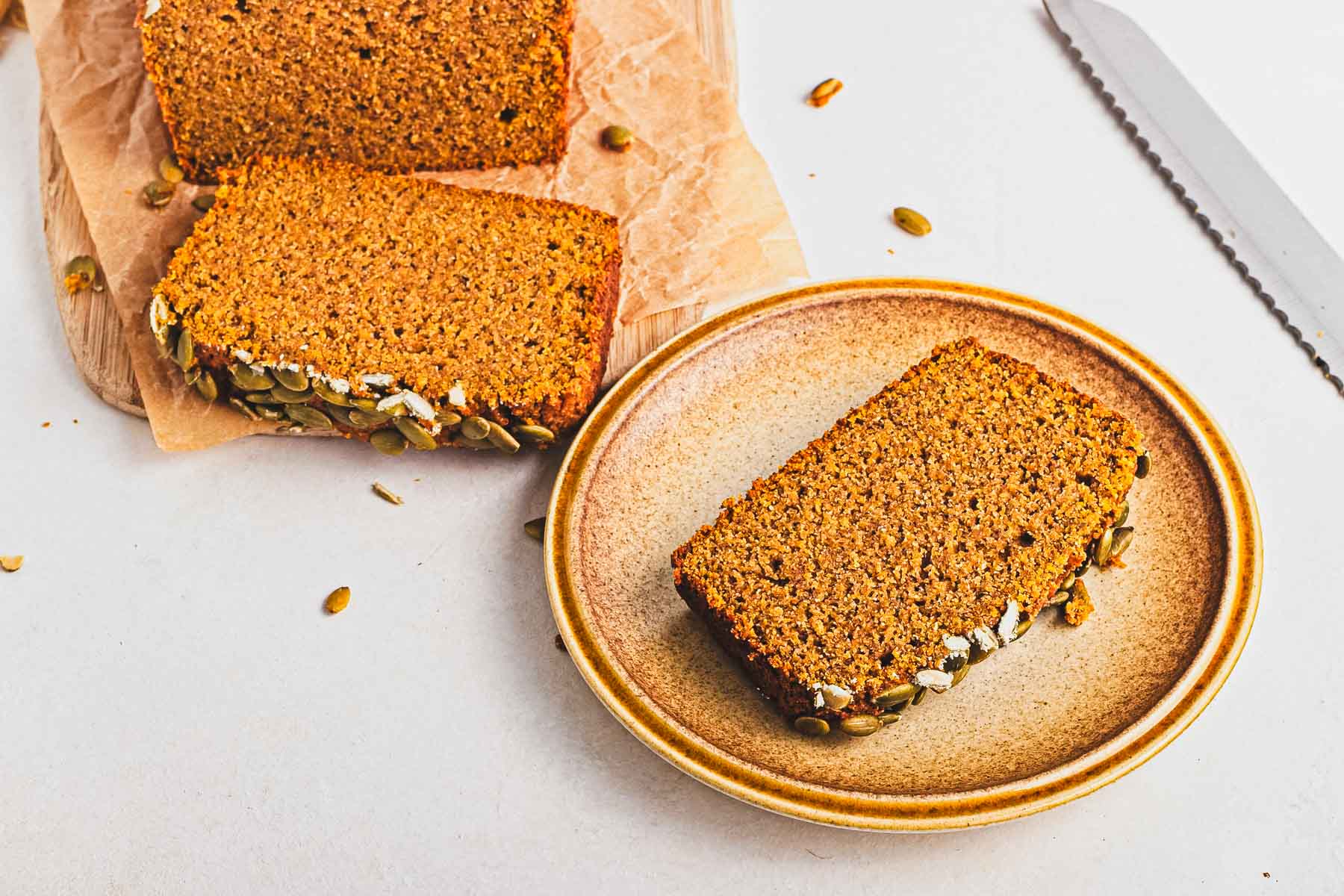 A slice of brown bread with seeds on a plate, next to more slices on a wooden board and a bread knife.