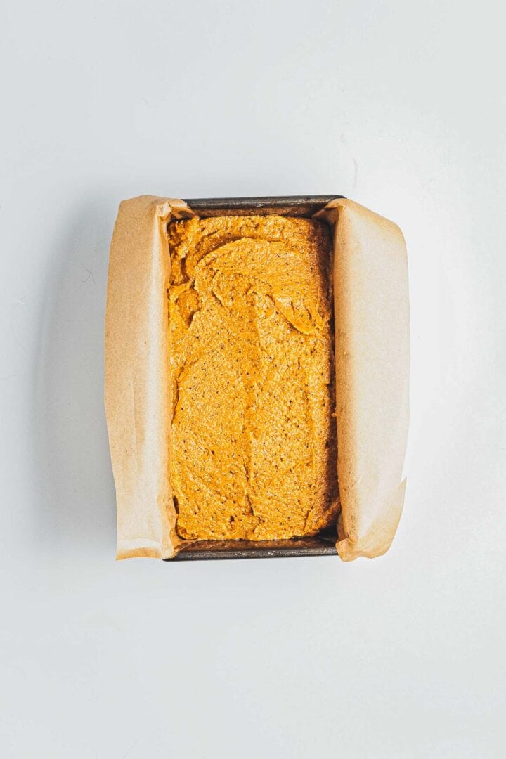 A loaf pan lined with parchment paper filled with pumpkin bread batter, ready to be baked.