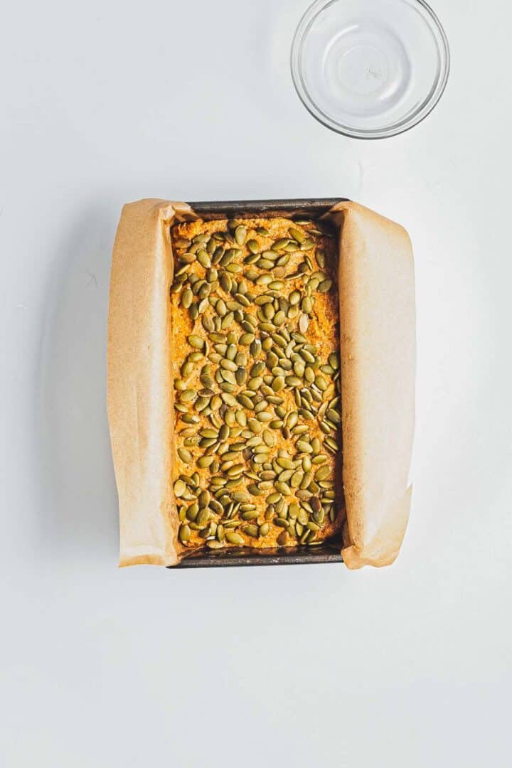 A loaf pan with pumpkin seed-topped bread batter lined with parchment paper, next to an empty glass bowl.