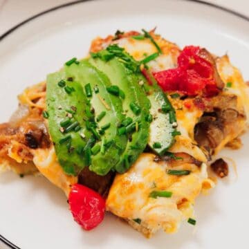 A serving of enchiladas topped with melted cheese, avocado slices, chives, and red salsa on a white plate.