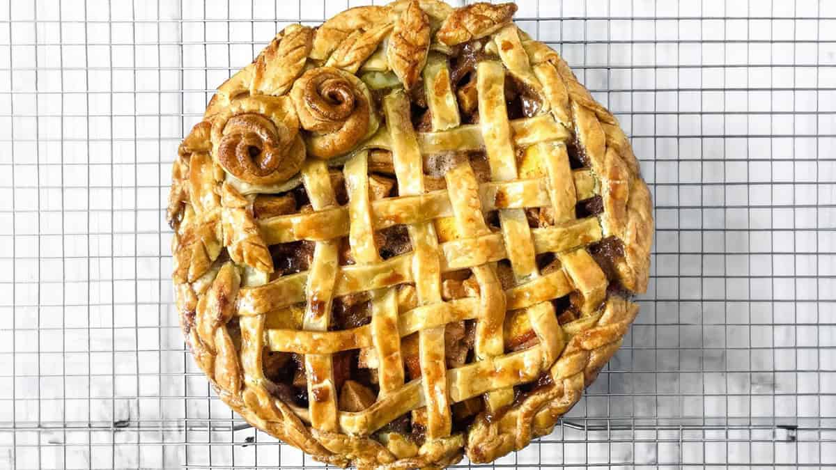 A pie with a golden-brown lattice crust and dough swirls rests on a cooling rack.