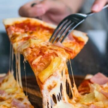 A close-up of a hand using a fork to lift a slice of pizza with melted cheese, pineapple, and ham from a wooden tray. Other pizza slices are visible, and a plate is in the background.