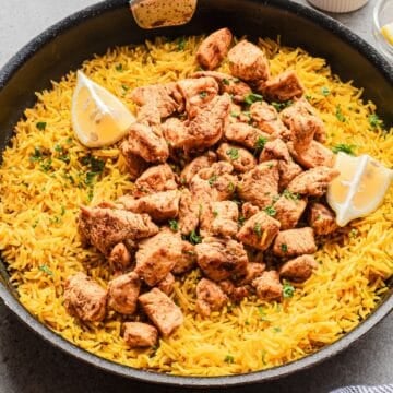 A pan of Chicken Shawarma Rice with White Sauce, topped with seasoned chicken pieces and garnished with lemon wedges and herbs.