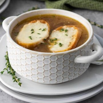 A white bowl filled with French onion soup, topped with two slices of bread covered in melted cheese and sprinkled with herbs, sits on stacked white plates next to a spoon and sprigs of fresh thyme.