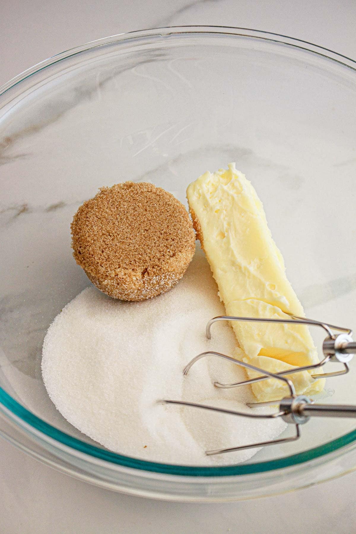 A glass bowl with brown sugar, white sugar, butter, and a hand mixer on a white countertop.