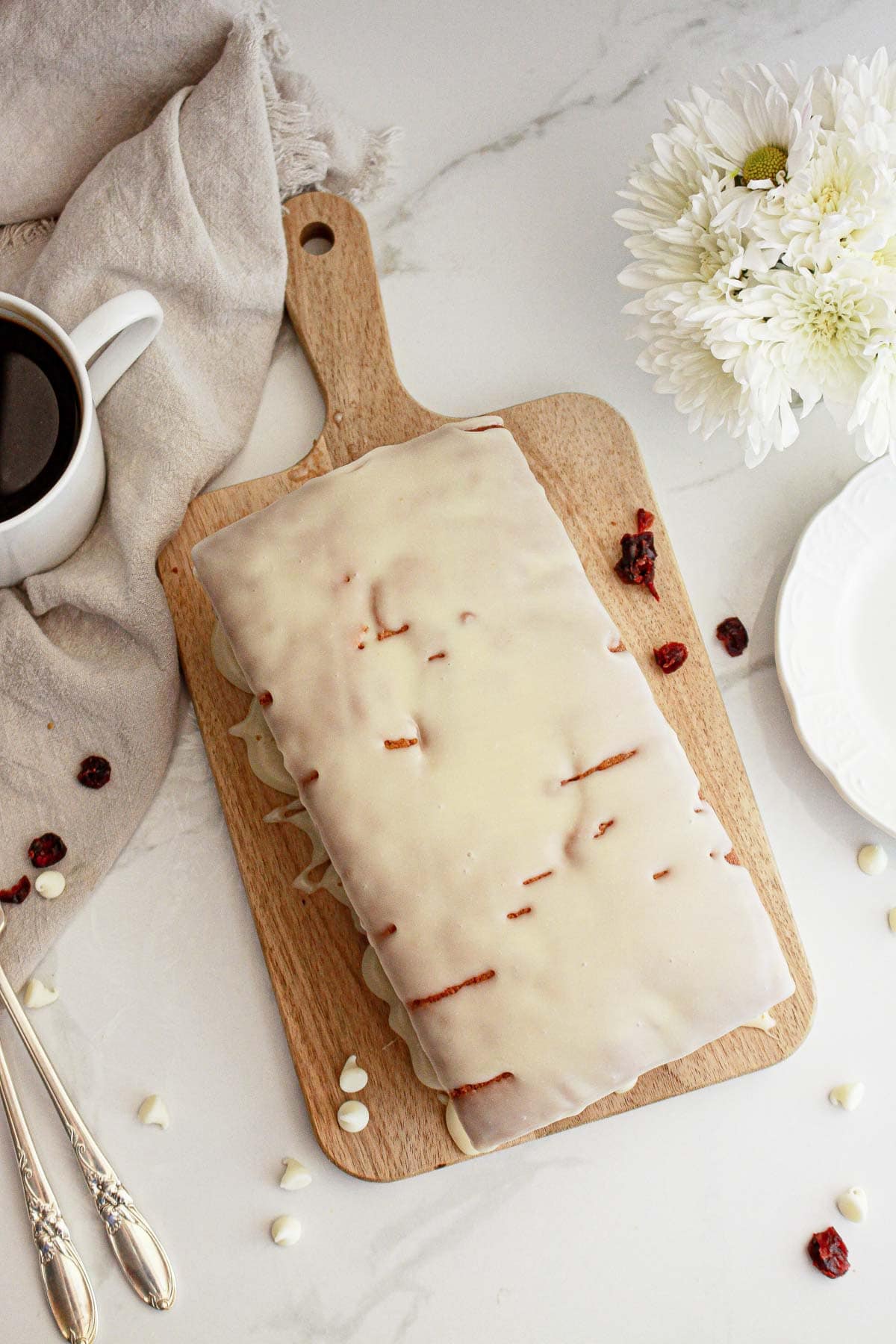 Rectangular iced loaf cake on a wooden board with coffee, white flowers, and scattered white chocolate chips.