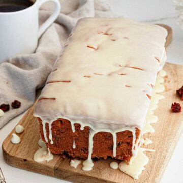 Cranberry White Chocolate Bread loaf with white glaze on a wooden board, next to a cup of coffee, forks, and dried fruit.