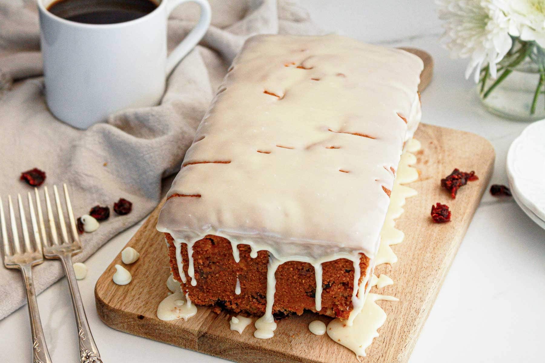 Iced loaf cake with white glaze on a wooden board, next to forks, a coffee cup, and white flowers.