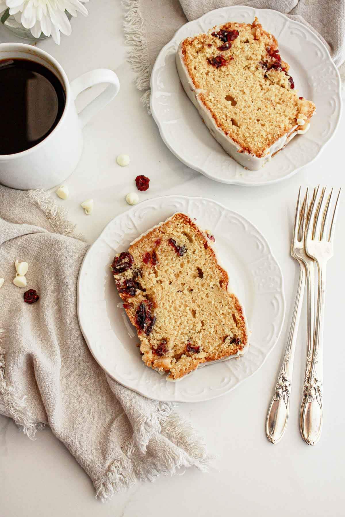 Two slices of cranberry bread on white plates with a cup of black coffee and two silver forks on the side.