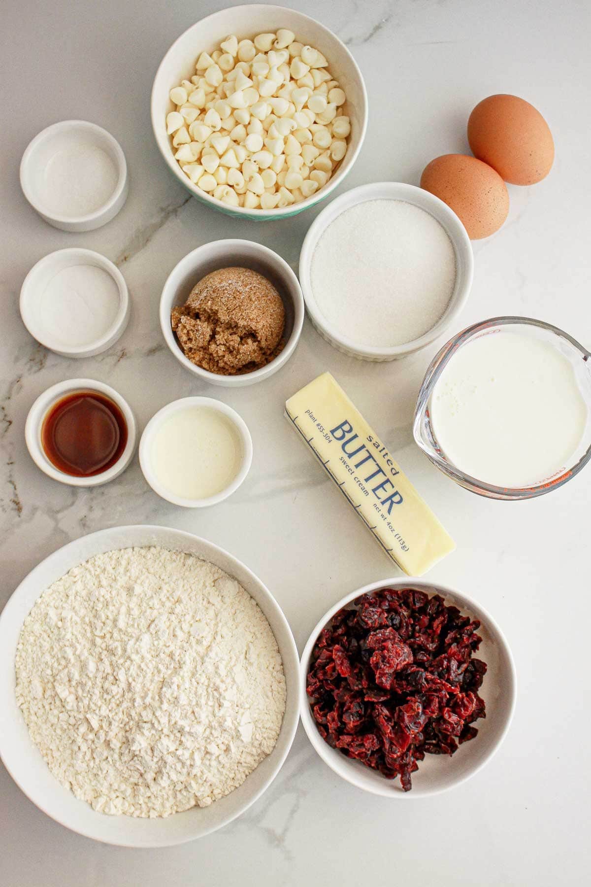 Bowls of white chocolate chips, dried cranberries, flour, sugar, brown sugar, eggs, and butter on a counter.
