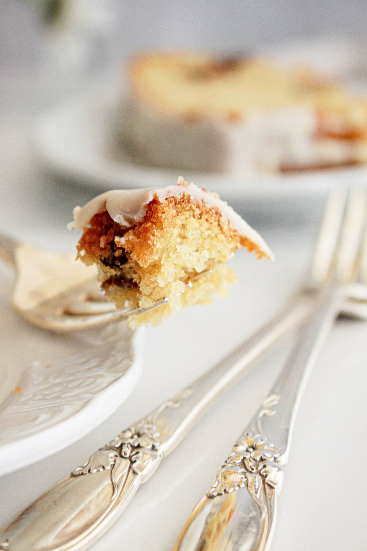 A fork holds a bite of iced cake, with a plate and utensils blurred in the background.