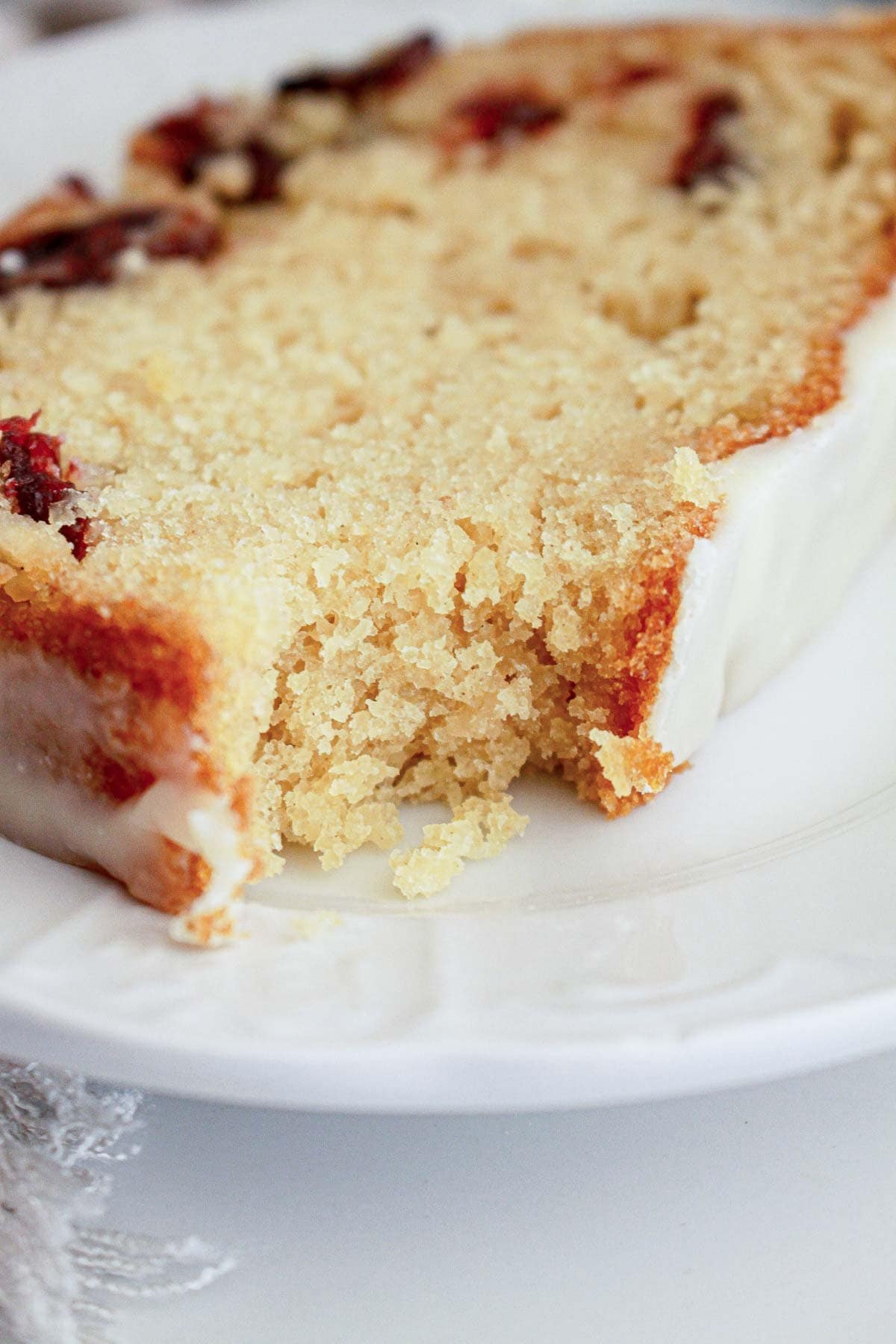 A close-up of a slice of glazed, moist loaf cake with bits of fruit on a white plate.