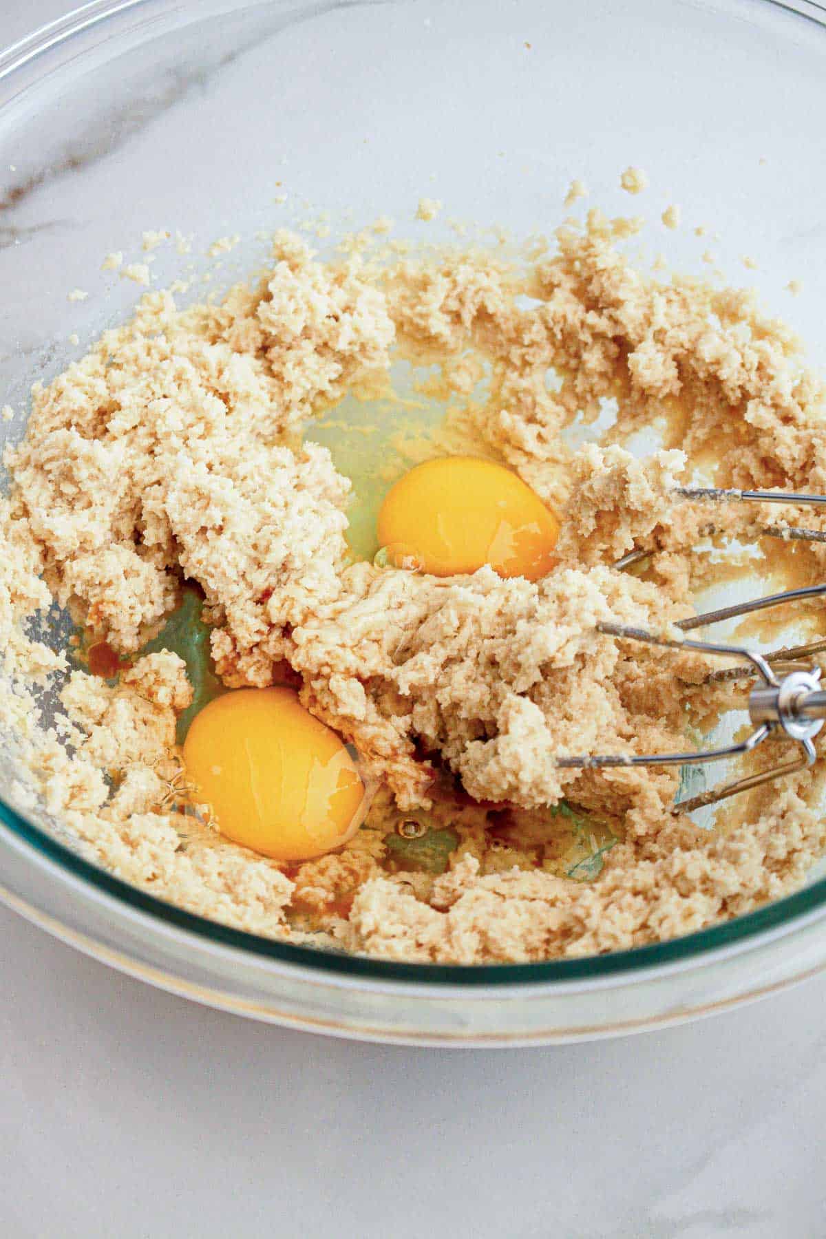 A glass bowl with cookie dough, two cracked eggs, and a hand mixer on a white surface.