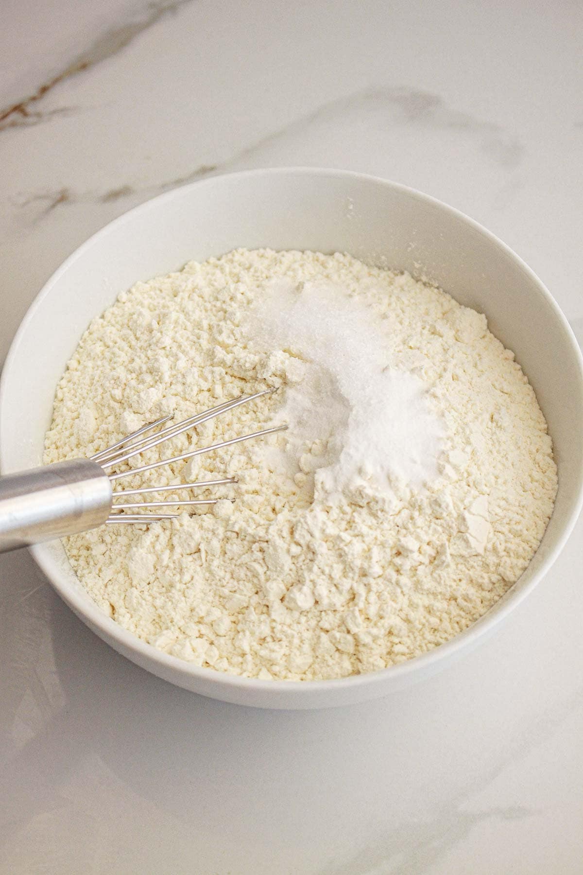A metal whisk in a bowl of flour and sugar on a white countertop.
