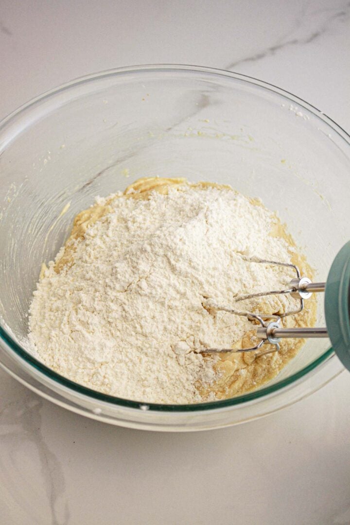 A glass bowl with flour and dough mixture, and an electric mixer on a marble countertop.