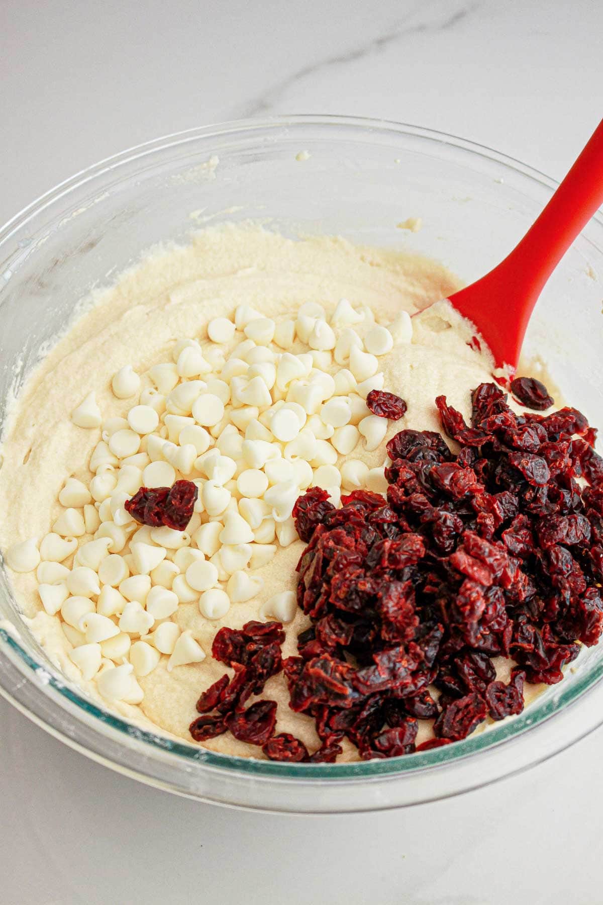 A mixing bowl with cookie dough, white chocolate chips, dried cranberries, and a red spatula.