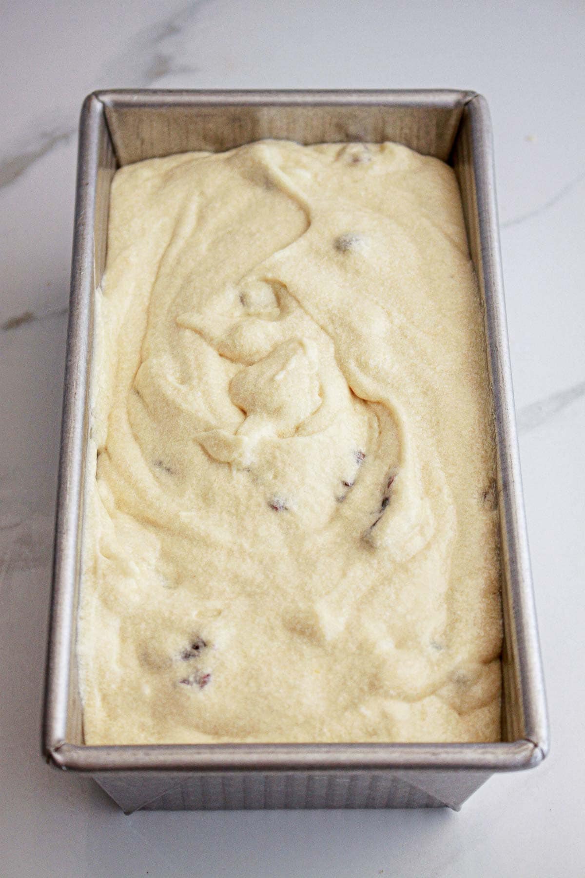 A metal loaf pan filled with unbaked batter containing small pieces, on a marble surface.