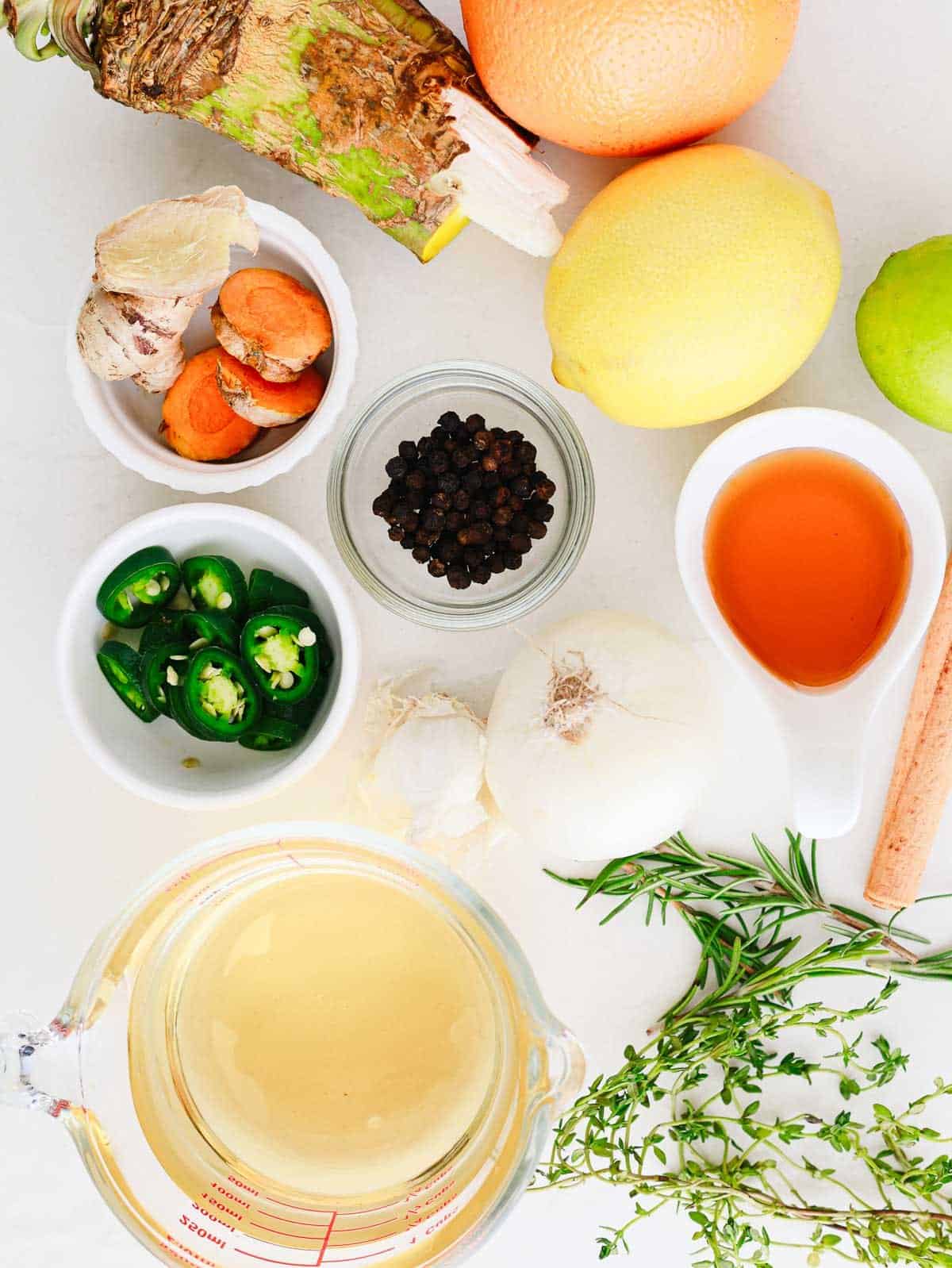 Assorted fresh herbs, spices, citrus fruits, peppers, and broth displayed on a white surface.