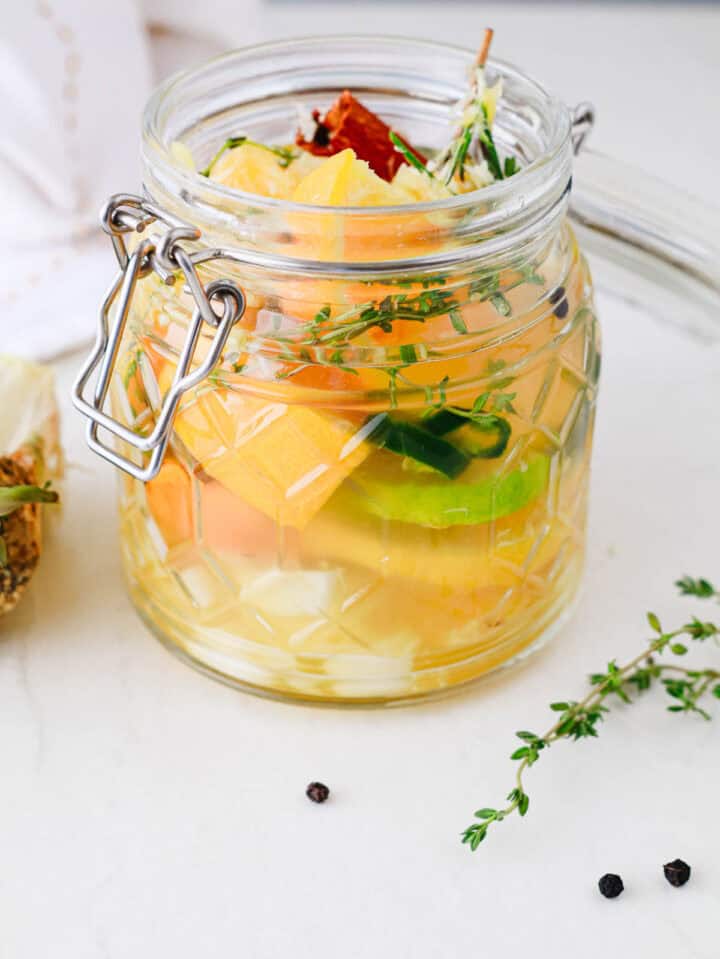 A glass jar of pickled vegetables and herbs sits open on a white surface with peppercorns and thyme nearby.