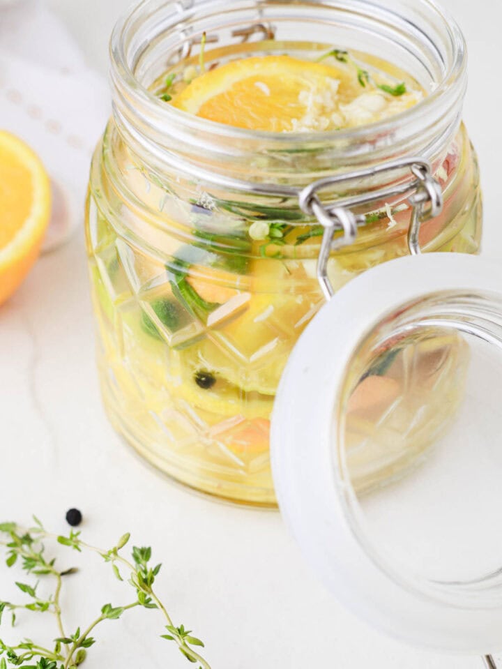 A glass jar with sliced citrus, herbs, and vegetables in liquid, with the lid open on a white surface.