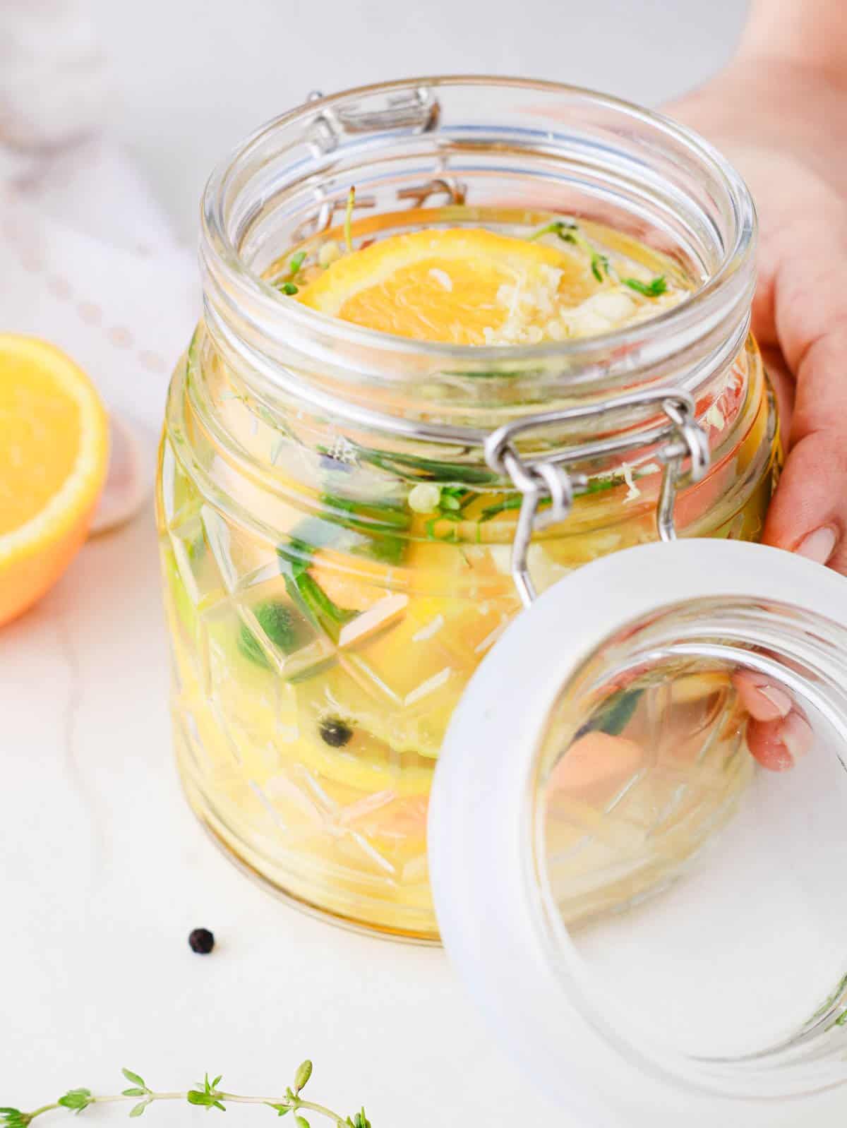A hand holding an open jar with citrus slices, herbs, and vegetables in liquid on a white surface.