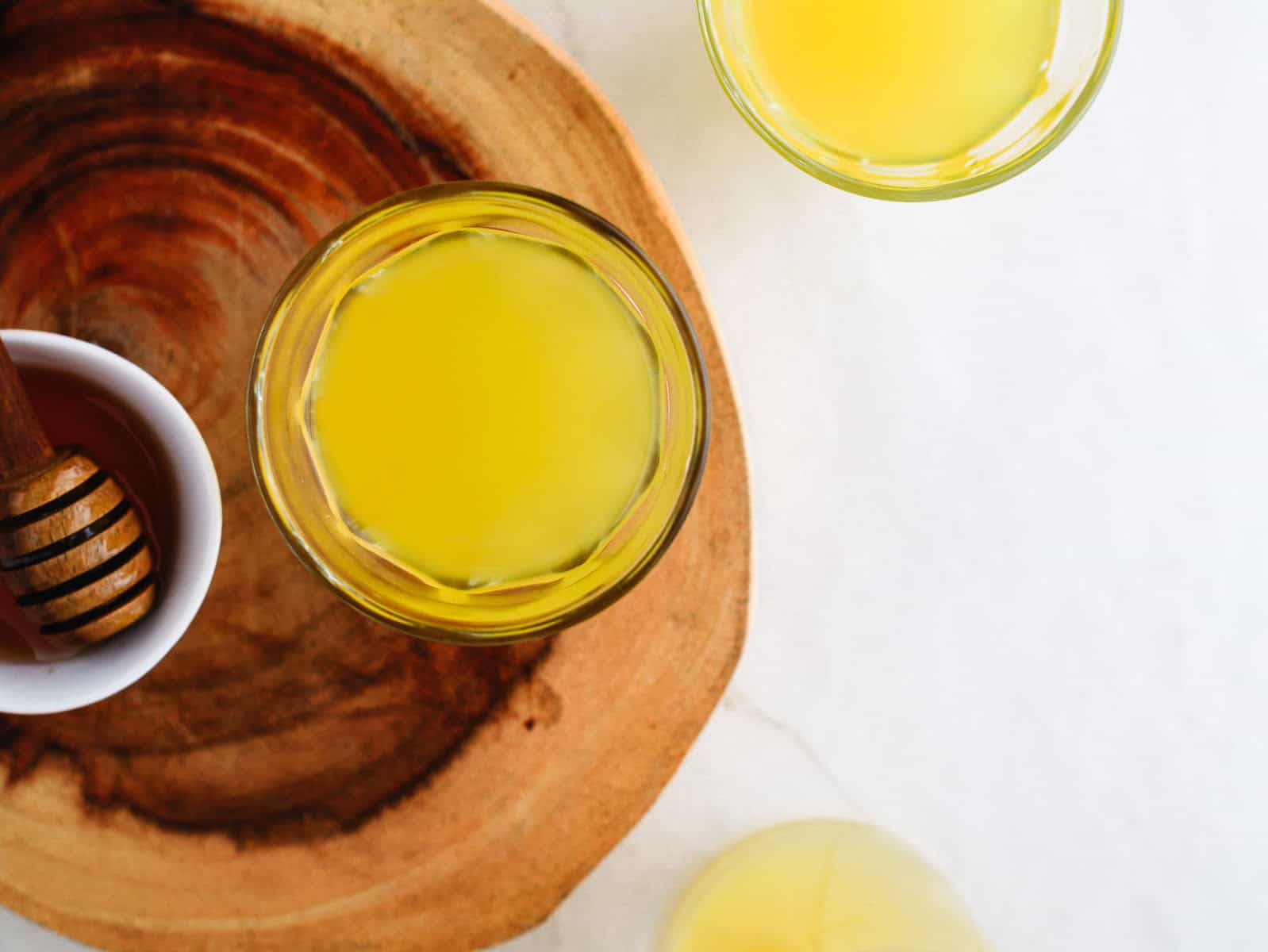 Glasses of yellow juice on a wooden tray with a bowl of honey and a honey dipper nearby.