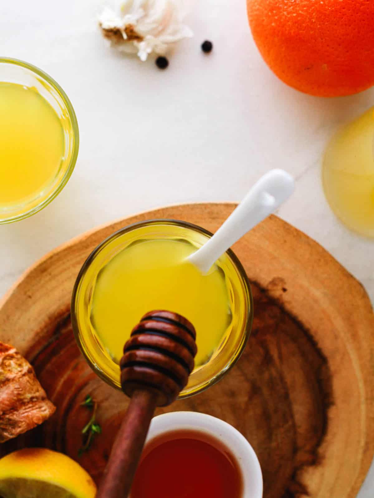 A glass of yellow liquid with a white spoon, honey dipper, and fresh ingredients on a wooden board.