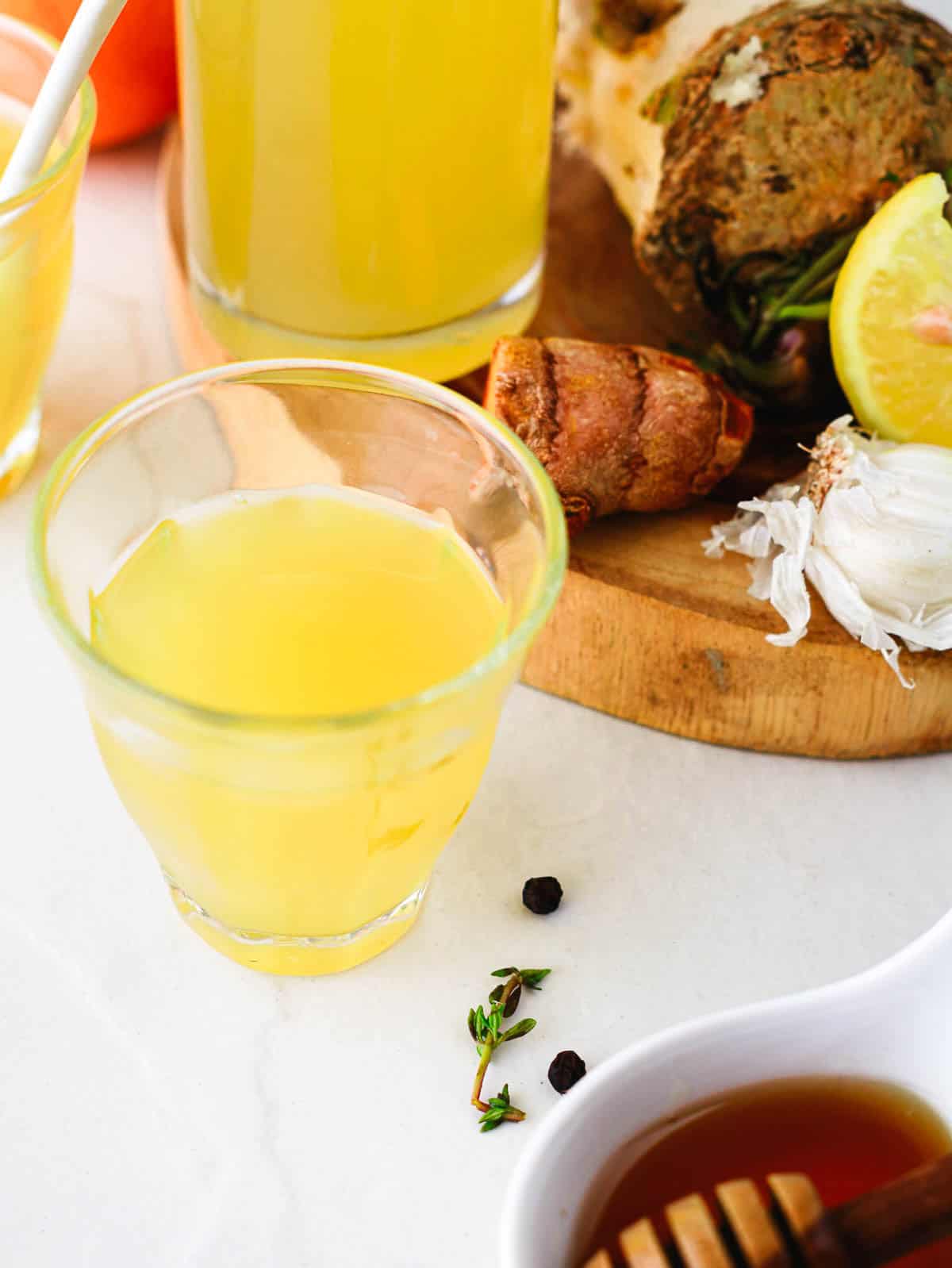 A glass of yellow juice on a table with herbs, spices, lemon, and honey nearby.